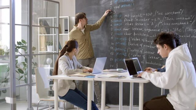 Young male professor of information technologies explaining material written on blackboard to small group of teenage students sitting at desks with laptops
