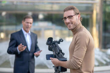 Smiling fair-haired operator filming mature male reporter in front of fountain