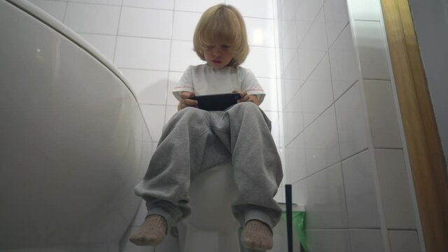 Handsome blond 5-aged boy playing game or watching cartoons on his mobile phone while sitting on the toilet bowl.