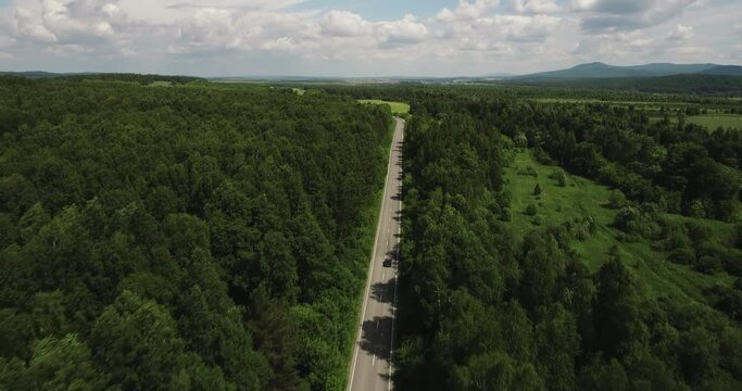 One dark car driving alone / travel on empty straight asphalt road freeway through dense forest corridor at summer sunny day. Aerial drone wide view.