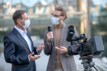 Male journalist interviewing mature male in front of camera, both wearing masks