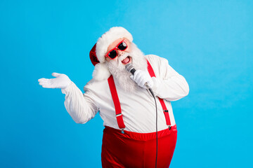 Fototapeta na wymiar Photo of old man grey beard hold microphone sing song raise palm wear santa claus x-mas costume cap sunglass suspenders white shirt gloves isolated blue color background