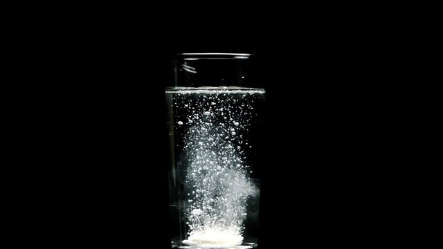 Effervescent or carbon tablet dissolves with bubbles in transparent glass with pure water, slow motion, isolated black background. Vitamins C pills against flu, medicines, aid, energy concept