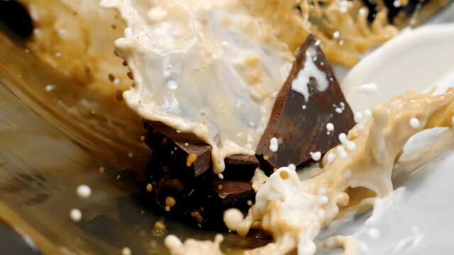 Dark chocolate stack in cocoa powder covered by splashing waves of white milk cream and dark coffee hot cocoa drink in slow motion on black background. Chocolate flavors, handmade chocolate concept
