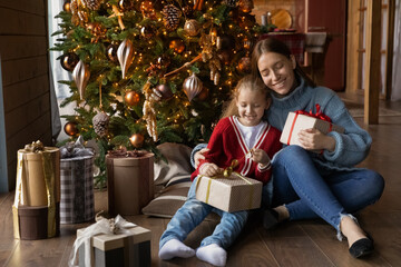 Obraz na płótnie Canvas Happy young Caucasian mom and small daughter sit near decorated Christmas tree unpack holiday presents together. Smiling mother and little girl child open New Year gift in cozy home living room.