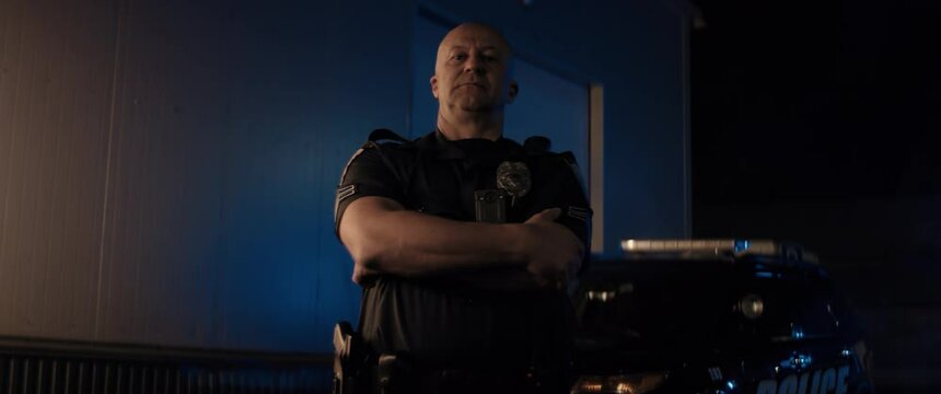 Caucasian male police officer posing against police car with flashing lights at night. Shot on RED cinema camera with 2x Anamorphic lens