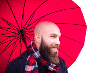 bald bearded man wearing a scarf and warm winter clothes holding a red umbrella as a background.smiling young adult on a white scenario in a studio shooting. concept about joy, happiness and beauty