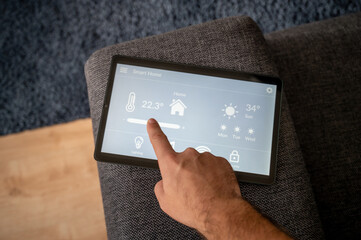 modern smart home control app with flat interface on black tablet display lies on the back of a sofa in front of a blue carpet a man controls the temperature of the heating