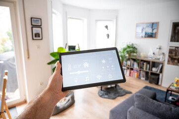 modern smart home controll app with flat interface on black tablet display. Men holding Tablet PC. Close-up in front of a blurry living room