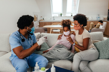 Medical worker is in protective work-wear and in home visit. Little girl is sick and her mother is helping her to use asthma pump