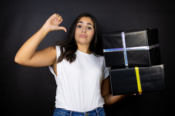 Young beautiful woman holding gifts over isolated black background looking confident with smile on...