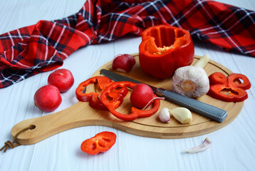Red bell pepper, garlic cloves, radish and a knife lie on a round cutting board.