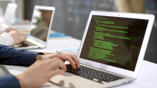 Closeup of laptop screen with program codes and male hands typing on keyboard while it teacher explaining material at blackboard