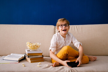 Blonde schoolboy boy plays video games, holds a gamepad, eats popcorn instead of learning lessons...