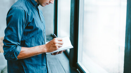 man making notes in a notebook, standing near the window.