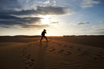 Fototapeta na wymiar Silhouette of a photographer shooting in the desert of Maspalomas, Gran Canaria at sunset. Sand dunes with footsteps.