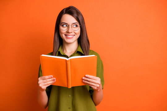 Photo portrait of curious female student wearing glasses keeping book looking at blank space isolated on vivid orange color background