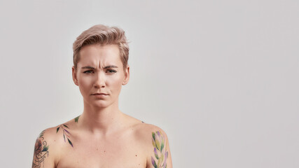 Still kind of miffed. Portrait of dissatisfied half naked tattooed woman with pierced nose and short hair frowning face at camera isolated over light background - obrazy, fototapety, plakaty