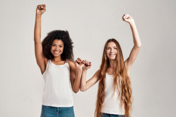 Portrait of two young diverse women, best friends smiling at camera while raising arm and making a...