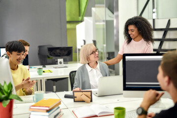 Aged woman, senior intern looking at her female colleague while completing first task at work, Friendly workers applauding, cheering new employee in the office