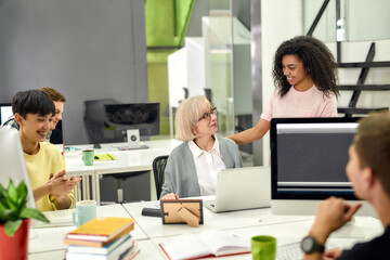 Obraz premium Aged woman, senior intern looking at her female colleague while completing first task at work, Friendly workers applauding, cheering new employee in the office