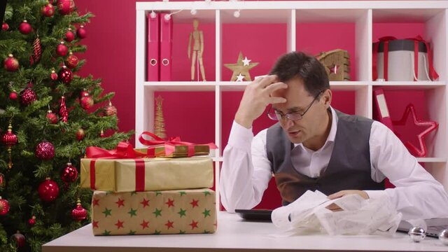 worried 40s businessman counts corporate holiday expenses on calculator and desperately grabs his head at Christmas decorated office. Festive depression and bankruptcy concept