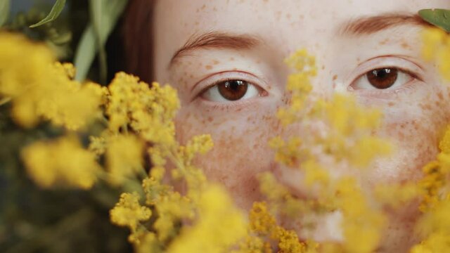 Close up tracking shot of redhead girl with freckles and brown eyes holding bouquet of mimosa flowers in front of her face and looking at camera