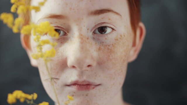 Close up shot of young beautiful woman with short red hair and freckles holding yellow wildflower in front of her face and looking at camera while posing against black studio background