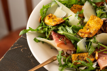 Light autumn salad with arugula, baked pumpkin, prosciutto and young parmesan, a plate of salad on a wooden table