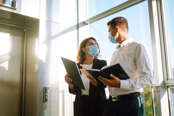 Two colleagues in protective face masks in modern office discussing together work issues standing near elevator. Business People back at work in office after quarantine. Covid-19.