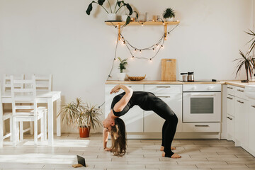 Woman of European appearance doing yoga in the kitchen. Sports at home online. A woman with long dark hair stands in a difficult yoga pose.