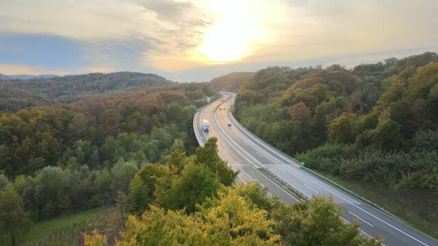 Aerial view of traffic highway through a green forest tree during colorful sunset autumn season. Green energy eco electric car no pollution.