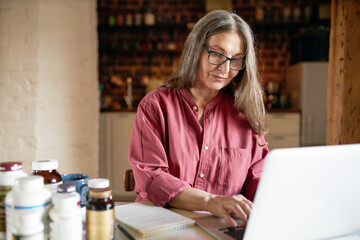 People, health, diet and wellbeing concept. Concentrated gray haired mature woman nutritionist typing on laptop, working on nutrition article, sitting at desk with bottles of food supplements