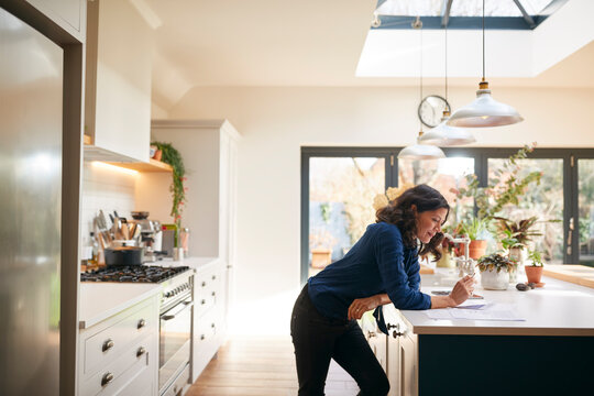 Mature Woman Reviewing And Signing Domestic Finances And Investment Paperwork In Kitchen At Home