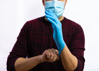 Caucasian man with a red and black sweater and surgical mask, on a white background he puts a blue latex glove on his hand. His eyes cannot be seen