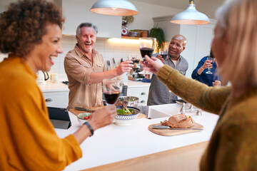 Group Of Mature Friends Making A Toast As They Meet At Home For Meal And Serve Food In Kitchen