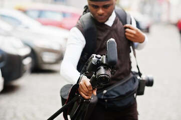 Young professional african american videographer holding professional camera with tripod pro equipment. Afro cameraman wearing black duraq making a videos.