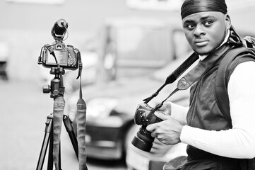 Young professional african american videographer holding professional camera with tripod pro equipment. Afro cameraman wearing black duraq making a videos.