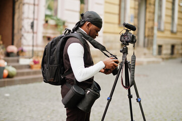 Young professional african american videographer holding professional camera with tripod pro equipment. Afro cameraman wearing black duraq making a videos.