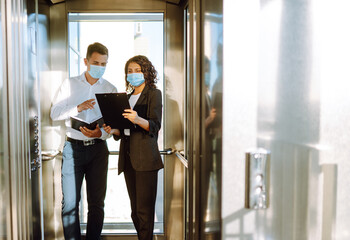 Business colleagues in medical face mask talking while standing in elevator at modern office. Boss...