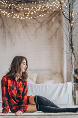 Obraz premium Pretty brunette girl sitting on the floor in red shirt and warm socks with christmas tree behind.