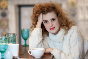 A beautiful girl in a white knitted sweater sits thoughtfully in a cafe. Inside