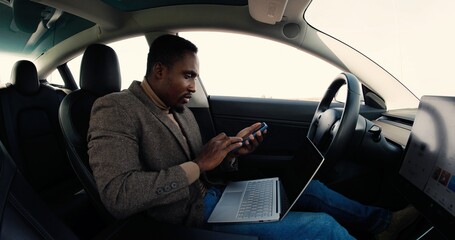 Concentrated young African American male sitting in modern electric car and browsing on computer. Handsome man holds smartphone and typing on laptop while traveling in autopilot vehicle. Tech concept