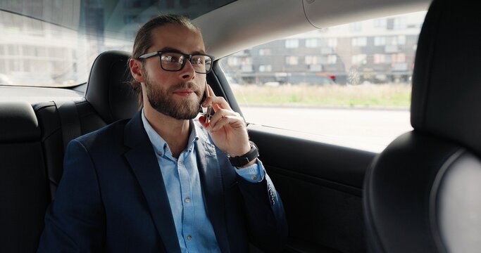 Portrait of handsome man on back seat in electric automobile speaking on mobile phone. Young Caucasian male professional sitting in automated car and talking on smartphone. Work concept