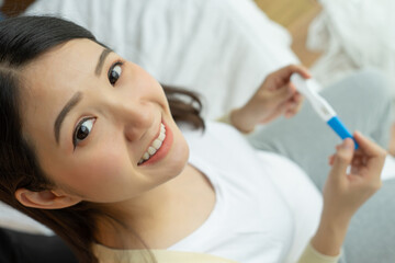 Happy woman sitting on bed looking at camera hand holding pregnancy test stick at home.