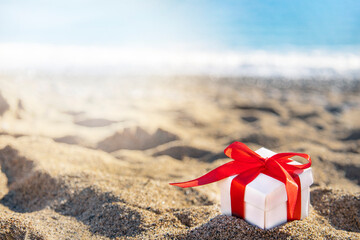 White gift box with a red ribbon standing on the sand with blue sea and sun on background. Holidays...