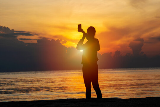 Silhouette of young woman using smartphone taking a selfie post on social media feeling relax over the beach with sunrise in the morning. Travel holidays vacation on nature sea concept.