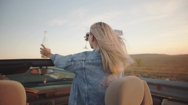 A Young Woman Rides Standing In The Front Seat Of A Cabriolet. She Has An Open Champagne Bottle In One Hand And A Wine Glass In The Other. She Takes A Sip From Her Glass And Holds It Up. Her Blond