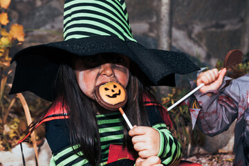 Fototapeta premium Girl dressed as witch eating candy celebrating Halloween in the garden next to Jack O Lantern