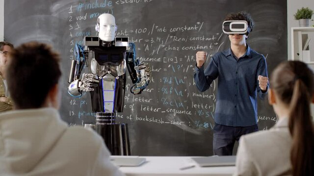 Panning footage of humanoid robot imitating movements of mixed race student wearing VR glasses standing at blackboard in front of teacher and classmates in equipped classroom